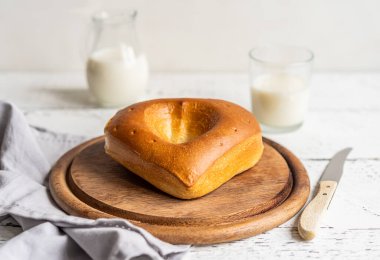 Tahta kesme tahtasındaki kalp şeklinde süt ekmeği. Kavanozda süt ve cam bir yana, gri peçete.