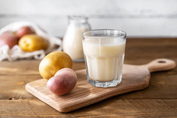Картофельное молоко в стекле и молочная банка на доске с сырым картофелем в струнном мешке. Деревянный фон. Сельский стиль — стоковое фото