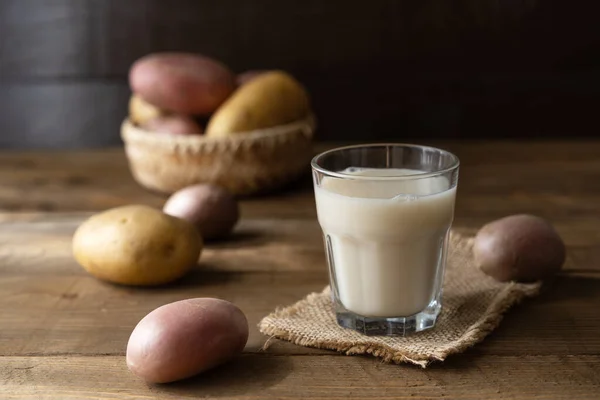 Веганское картофельное молоко в стекле и молочной банке с сырым картофелем на деревянном фоне в деревенском стиле — стоковое фото