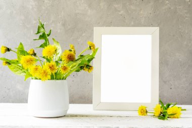 Mock up creation. White frame on white wooden table with grey concrete background and dandelions in vase