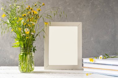 Yaratılışı taklit et. Beyaz çerçeve beyaz ahşap masa üzerinde gri beton arka plan, kitaplar, defter ve yabani çiçekler