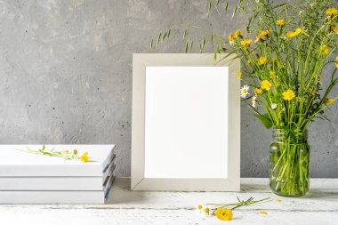 Yaratılışı taklit et. Beyaz çerçeve beyaz ahşap masa üzerinde gri beton arka plan, kitaplar, defter ve yabani çiçekler