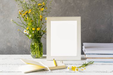 Yaratılışı taklit et. Beyaz çerçeveli ahşap masa, gri beton arka plan, kitaplar, defter, kalem ve yabani çiçekler.