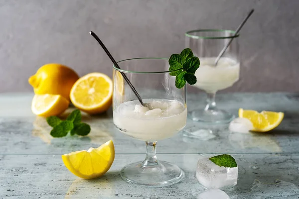 Σορμπέ λεμόνι σε δύο ποτήρια με λεμόνια, παγάκια, κουταλάκια τσαγιού, φύλλα δυόσμου σε γκρι φόντο — Φωτογραφία Αρχείου