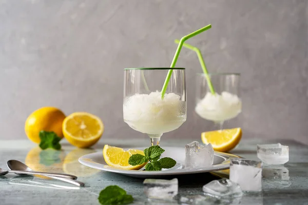 Lemon sorbet in two glasses on plate with lemons, ice cubes, mint leaves and drinking straws on grey background