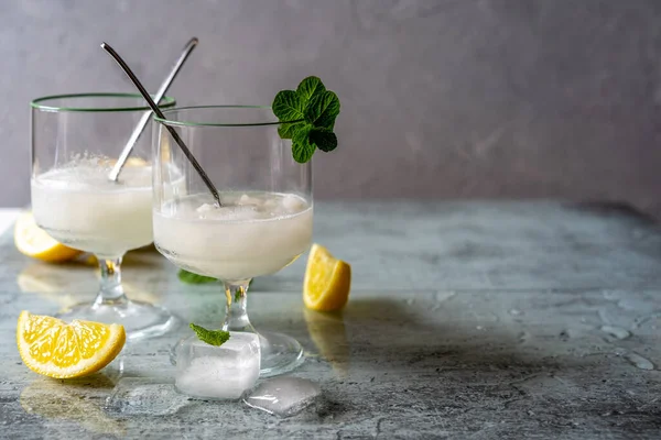 Lemon sorbet in two glasses with lemons, ice cubes, tea spoons, mint leaves on grey background with copy space