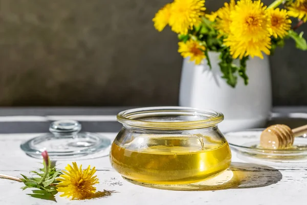 Karahindiba balı, kavanozda, beyaz ahşap masada çiçekler ve cam tepside bal dolu kaşık.