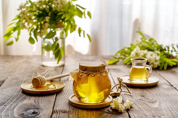 Acacia honey in glass jar with wooden dipper and flowers aside on wooden background