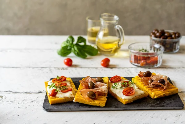 Üzerine strakino peyniri, domates, otlar, benek ve zeytin konmuş ızgara polenta sosu. Beyaz ahşap arka plan. Beyaz şarap arkada. Uzayı kopyala İtalyan antipasto kavramı.