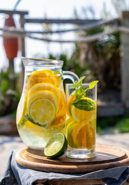 Infused water with lime and oranges in transparent jug and glass on the beach. Selective focus. Vertical banner