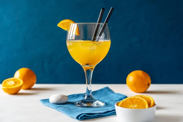 Yellow bird cocktail with rum, orange and lime juice in glass with slice of orange and two straws, fruits, blue napkin on white table. Dark blue background. Mediterranean or marine style. Copy space