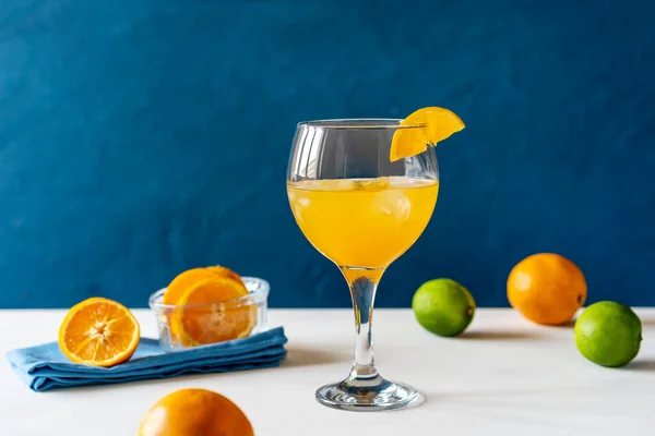 Yellow bird cocktail with rum, orange and lime juice in glass with slice of orange, fruits, blue napkin on white table. Dark blue background. Mediterranean or marine style. Copy space