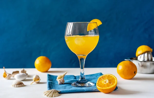 Yellow bird cocktail with rum, orange and lime juice in glass with slice of orange, fruits, juicer, shells on white table, napkin. Blue background. Mediterranean and marine style. Copy space