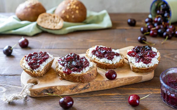 Kepekli ekmek, ricotta ve ahşap kesme tahtasının üzerinde ev yapımı kiraz reçeli kavanozda reçel, taze kiraz üzümü ve çörekle yapılan tostlar. Ahşap arka plan. Sağlıklı ve mevsimsel gıda konsepti.