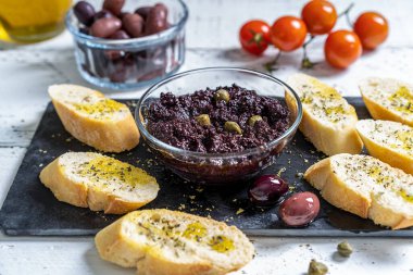 Homemade olive pate or tapenade in bowl, made with capers, black olives crushed and added with olive oil. Pate in bowl, ingredients, crostini with oil and oregano. White wooden background. 