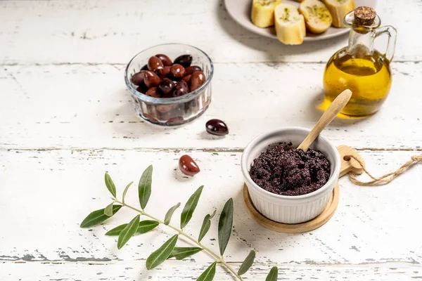 Homemade olive pate or tapenade made with capers, black olives crushed and added with olive oil. Pate in ramequin bowl, ingredients, crostini, olive branch aside. White wooden background. Copy space