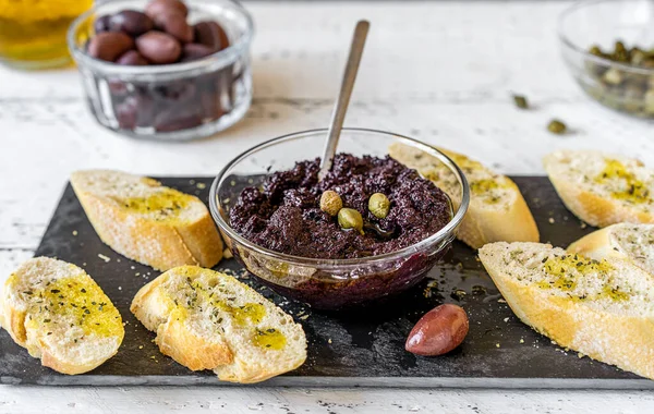 Ev yapımı zeytin ezmesi ya da kapari ya da zeytinyağıyla ezilmiş siyah zeytinlerden yapılmış tapenata. Tabakta börek, malzemeler, yağlı ve kekikli crostini. Beyaz tahta arkaplan. 