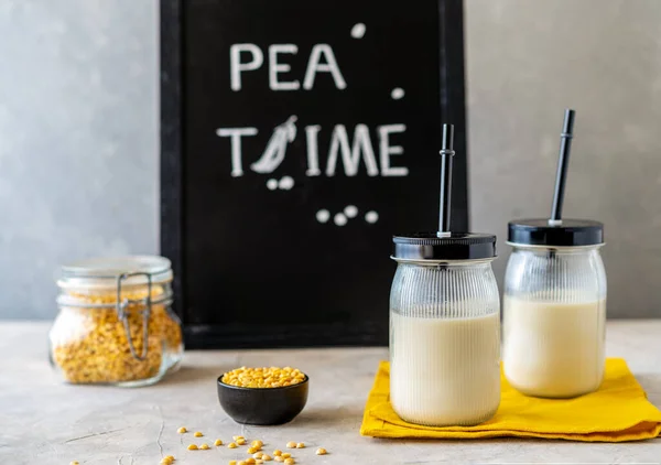 Vegan healthy plant pea milk in jars with drinking straws, yellow pea grains, napkin and mockup place. Concrete background.