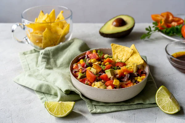 Cowboy caviar is traditional Mexican vegetable salad in bowl made with avocado, black beans, bell pepper, corn, coriander, lime juice and dip. Concrete background, green napkin, nachos