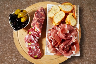 Siyah ve beyaz zeytinli ahşap kesme tahtası, zeytinyağlı kızarmış ekmek, şam fıstıklı salam ve Sicilya ham jambonu.
