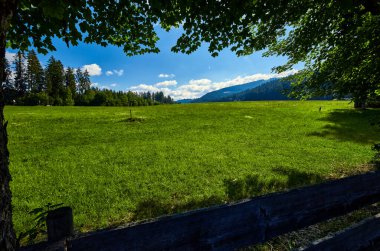 El değmemiş yeşil dağ manzarası, arka planda dağlar ve ağaçlar ve koreografik bulutlarla dolu mavi gökyüzü.