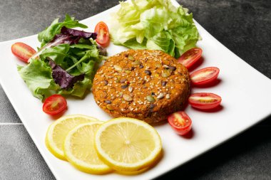 Lentils, legumes and various seeds burger, served on a salad, with cherry tomatoes and lemon slices