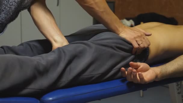 Braços de massagista fazendo rubdown cura de quadril para jovem atleta deitado na mesa de massagem. Mãos masculinas de massagista massajando de volta da coxa para desportista muscular em salão. Conceito de estilo de vida saudável — Vídeo de Stock