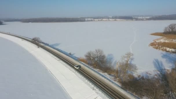 Foto aerea di auto che attraversa la strada coperta di neve vicino al lago ghiacciato. SUV bianco guida a rotta diga il giorno d'inverno. Volare sopra l'auto in movimento attraverso il ponte del fiume. Paesaggio panoramico. Vista dall'alto — Video Stock