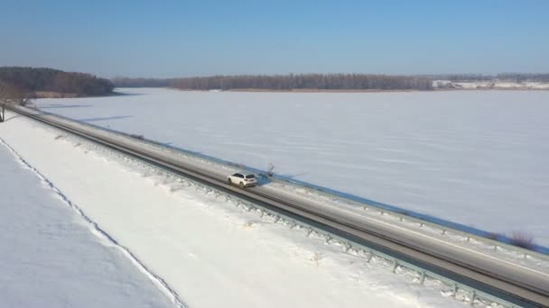 Flygfoto av bil ridning genom snötäckta väg nära frusen sjö. Vit SUV kör vid dammen rutt på vinterdagen. Flyger över auto rör sig genom bron över floden. Det natursköna landskapet. Ovanifrån — Stockvideo