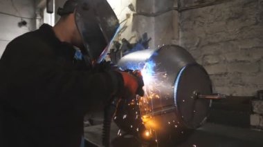 Metal fabrikasında koruyucu maske kaynak işçisi. Kaynakçı, endüstriyel üretimde iki çelik detay arasında ortak iş yapıyor. Adam metal parçalarını birbirine kaynaştırıyor.