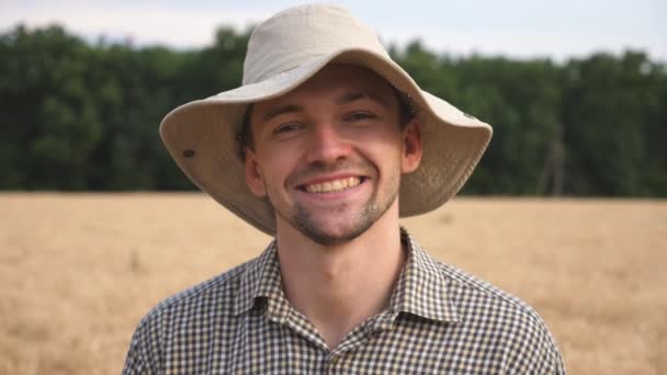 Primer plano de feliz granjero sonriente con sombrero mirando a la cámara contra el fondo borroso del campo de trigo. Retrato de un joven agrónomo guapo parado en el prado de cebada. Concepto agrícola — Vídeo de stock