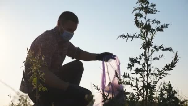 Άντρας οικολόγος ακτιβιστής με γάντια που μαζεύει σκουπίδια στην εξοχή. Νεαρός εθελοντής σε μάσκα καθαρό γρασίδι από απορρίμματα χαρτιού κοντά σε οδική διάσωση της φύσης. Έννοια του περιβαλλοντικού προβλήματος. Αργή κίνηση — Αρχείο Βίντεο