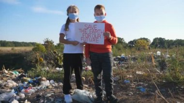 Küçük oğlan ve kız dünyayı kurtarmak için çevresel hareketin posterini tutuyorlar. Koruyucu maskeli küçük eko gönüllüleri kameraya bakıyor ve çöplerin arkasında duruyor. Kapat.