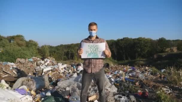 Mannelijke eco-activist in beschermend masker kijkt in camera houden in handen banner van milieubeweging. Jongeman tegen de achtergrond van dumping en het oprichten van een bord voor het redden van de natuur — Stockvideo