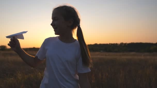 Boldog kislány, aki papírrepülővel szaladgál a füves mezőn. Vidám kis gyermek kocog játék sík között rét felett naplemente háttér. Aranyos gyerek játszik a természetben. A gyermekkori álom fogalma — Stock videók