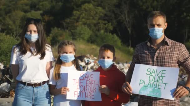 Młoda para z dwójką dzieci w medycznych maskach ochronnych stojąca na tle wysypiska śmieci. Rodzina eko-aktywistów trzymających w rękach plakaty ruchu ekologicznego dla ratowania planety — Wideo stockowe