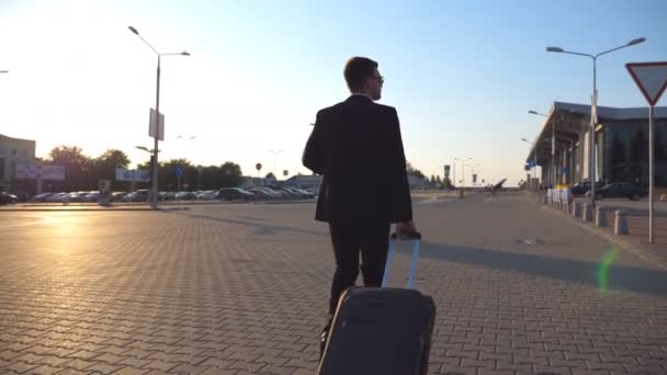 Dolly shot van zelfverzekerde jongeman in een formeel zwart pak lopen met zijn bagage op stedelijke straat. Succesvolle zakenman gaat naar de luchthaven terminal en trekt koffer op wielen bij zonsondergang. Sluiten. — Stockvideo
