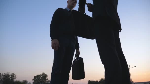 Dolly shot of young managers with briefcases bids farewell with each other and shaking hands at city on sunset. Two colleagues are leaving in different directions after meet outdoor. Low angle view — Vídeos de Stock