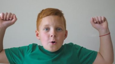 Sevimli, kızıl saçlı, çilli bir çocuk kameraya bakıyor ve el kaldırıyor. İçindeki başarıyı alkışlıyor. Yüzünde mutluluk ve sevinç duyguları olan mutlu genç bir çocuğun portresi. Yavaş çekim