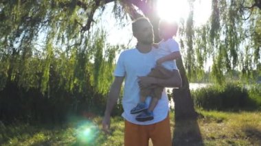 Genç baba oğlunun elinde tutan ve güneşli bir günde yaz Park'ta yürüyüş. Birlikte vakit ve doğanın keyfini mutlu bir aile. Arka plan, güzel manzara. Ağır çekim yakın çekim