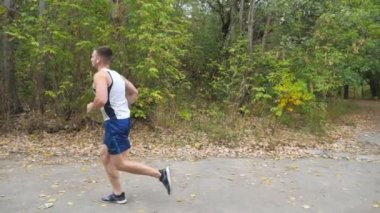 Kablosuz kulaklıklı güçlü genç sporcu ormanın yakınındaki patikada koşuyor. Sportif adam doğada yol boyunca koşuyor. Atletik adam sonbaharın başında egzersiz yapıyor. Sağlıklı aktif yaşam tarzı kavramı