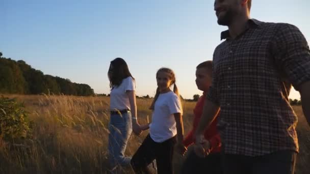 Happy family with two little children going among summer meadow and enjoying beautiful nature. Young parents with small kids walk through grass field holding hands of each other at sunset. Dolly shot — Stockvideo