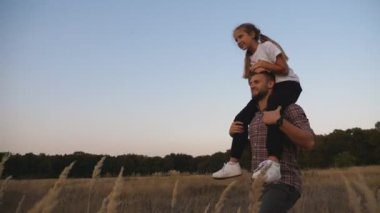 Genç baba, çayırda omuzlarında oturan tatlı küçük kızıyla yürüyor. Mutlu baba ve küçük çocuğu alacakaranlıkta çim tarlasında birlikte vakit geçiriyorlar. Aile mutluluğu kavramı