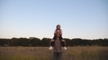 Genç baba, çayırda omuzlarında oturan tatlı küçük kızıyla yürüyor. Mutlu baba ve küçük çocuğu alacakaranlıkta çim tarlasında birlikte vakit geçiriyorlar. Aile mutluluğu kavramı