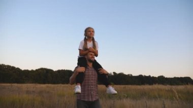 Genç baba, çim tarlasında omuzlarında oturan tatlı küçük kızıyla yürüyor. Küçük bir çocuk babasını işaret ediyor. Mutlu aile akşam karanlığında Meadow 'da birlikte vakit geçiriyor. Kapat.