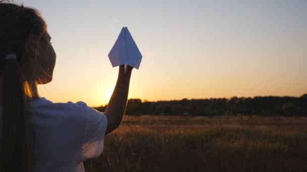 Happy small girl stand at grass field holding in hand a paper airplane. Little female child playing with toy plane at meadow over sunset background. Carefree kid having fun at nature. Childhood dream — Video Stock