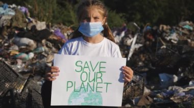 Tıbbi koruyucu maskeli küçük bir kız, el ele tutuşup, çevresel hareketlerin posterini tutan kameraya bakıyor. Küçük ekolojik aktivist, doğayı çöplerin arka planına karşı korumak için levha kaldırıyor.
