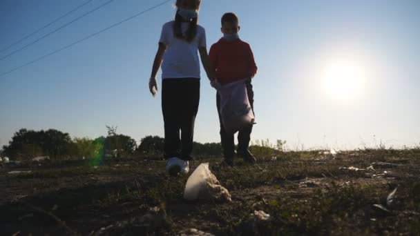 Small eco volunteers in masks cleaning lawn of trash near roadside. Brother holding bag while his sister in gloves throwing paper waste in bag. Little girl and boy help to saving nature. Close up — Stock Video