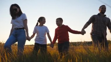 Happy parents with small kids walk through grass field holding hands of each other at sunset. Mother and father with two children going among summer meadow and enjoy to spending time together. Slow mo