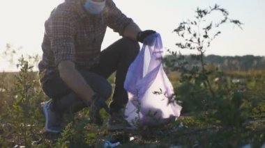 Young volunteer in mask clean lawn of paper waste near roadside saving nature. Male eco activist in gloves collecting trash in bag at countryside. Concept of environmental problem. Dolly shot
